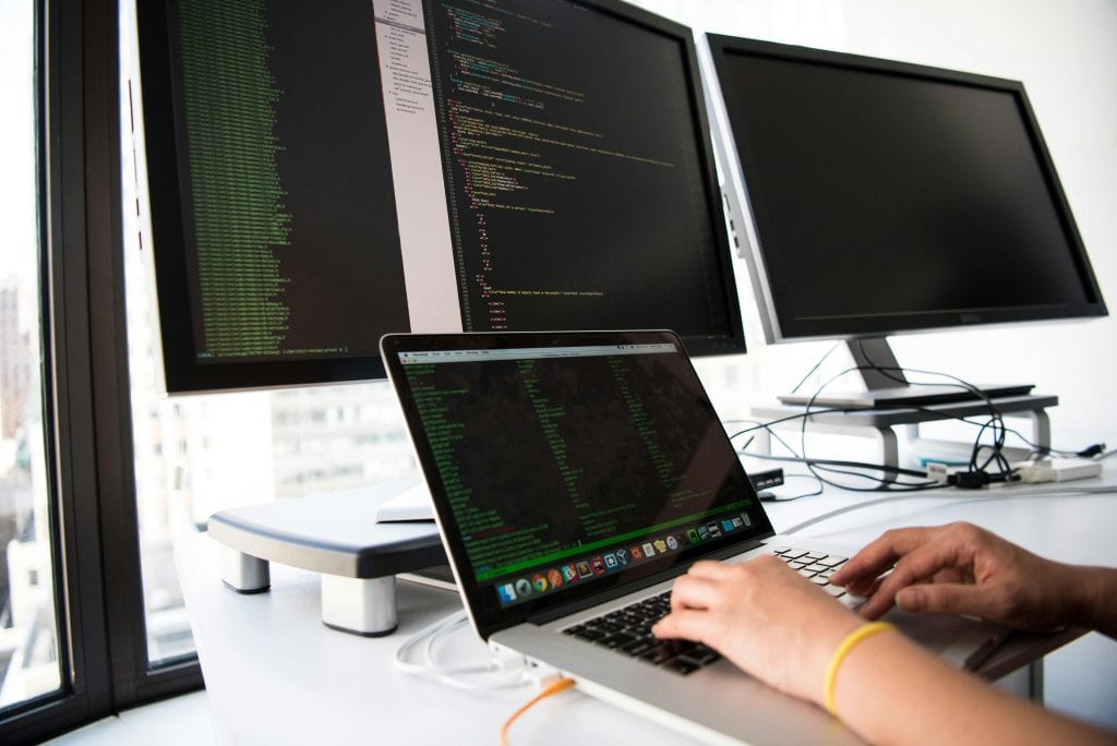 A man typing on a silver laptop and sitting in front of two monitors