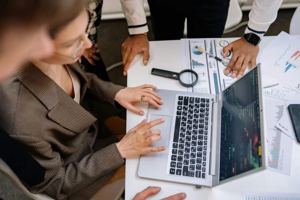A woman is using a laptop with her colleagues while paper documents, pen, and a magnifying glass are on the table