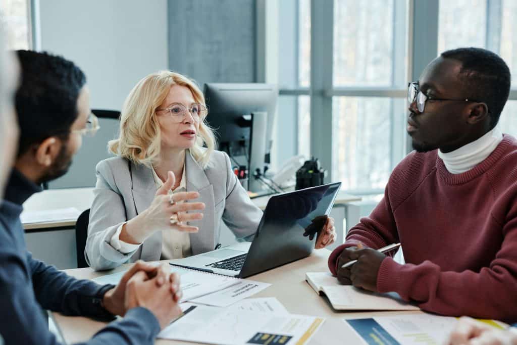 A woman in gray blazer is discussing with her colleagues while holding a laptop