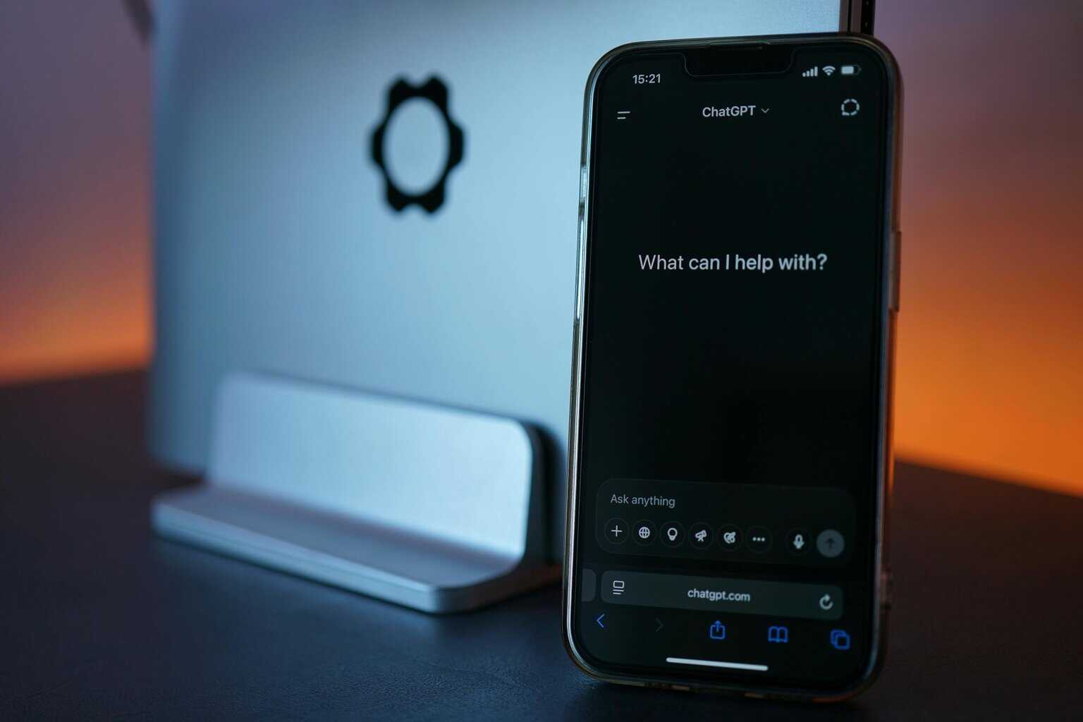 A phone showing ChatGPT's website on screen with a desktop monitor in the background