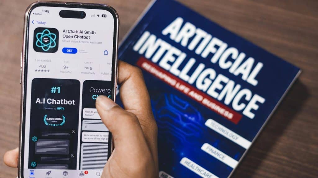 Hand holding a phone displaying an AI chatbot app beside an “Artificial Intelligence” book