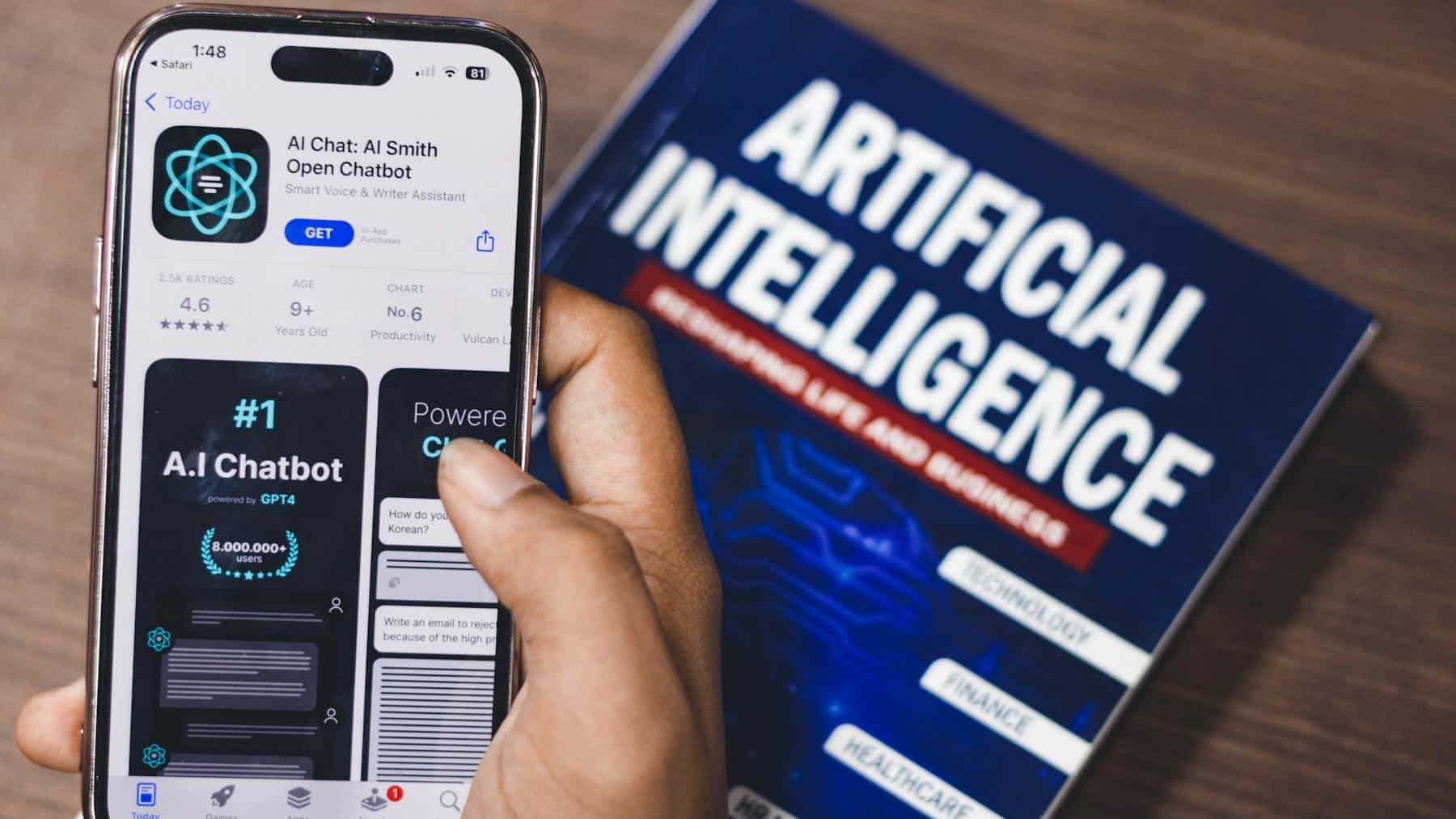 Hand holding a phone displaying an AI chatbot app beside an “Artificial Intelligence” book