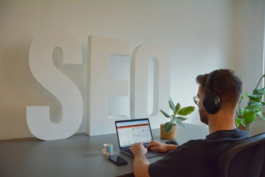 Man wearing headphones working on a laptop at a desk with giant “SEO” letters